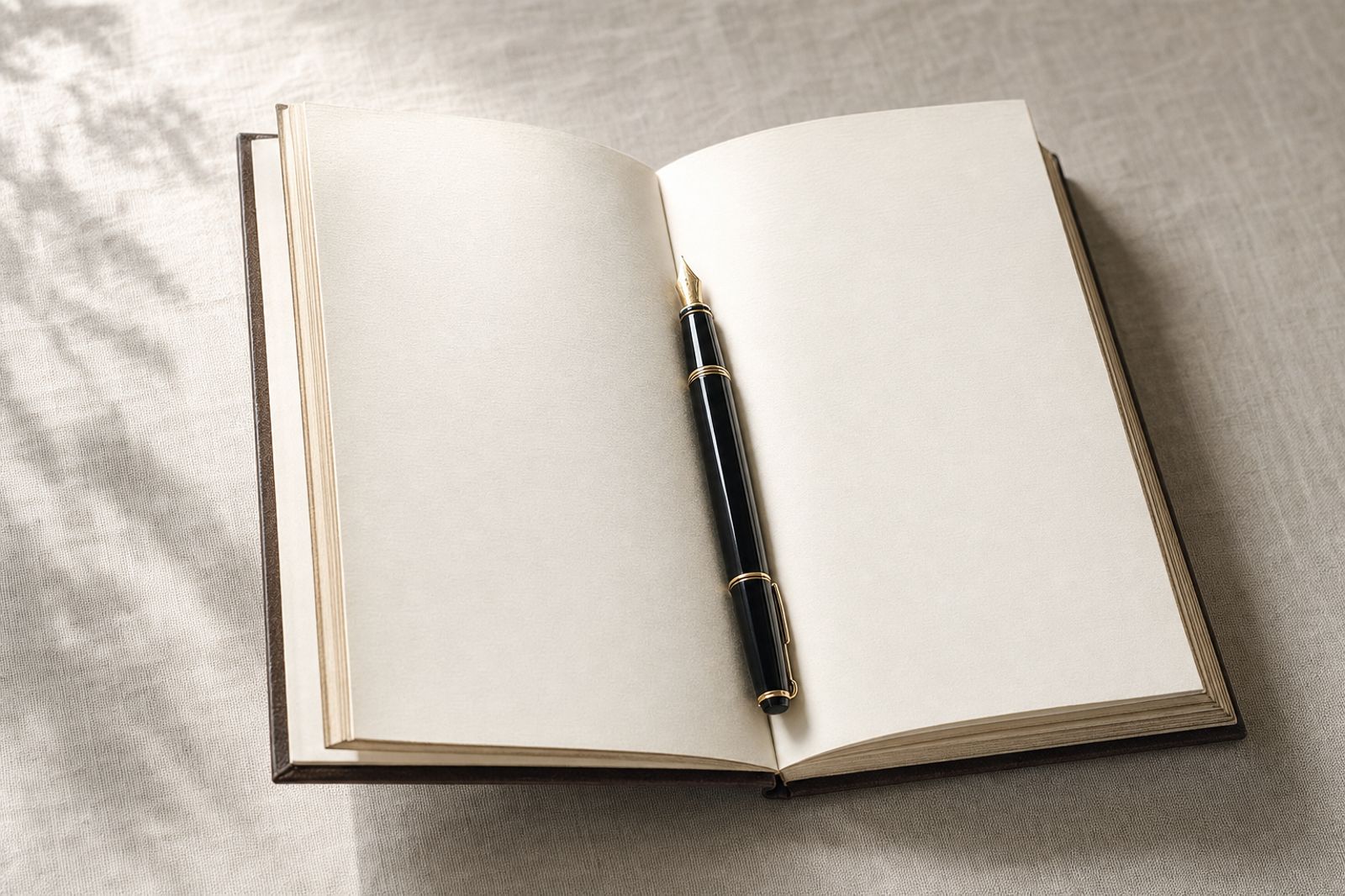 An open hardcover ledger on linen with a single fountain pen resting across the gutter, side light.